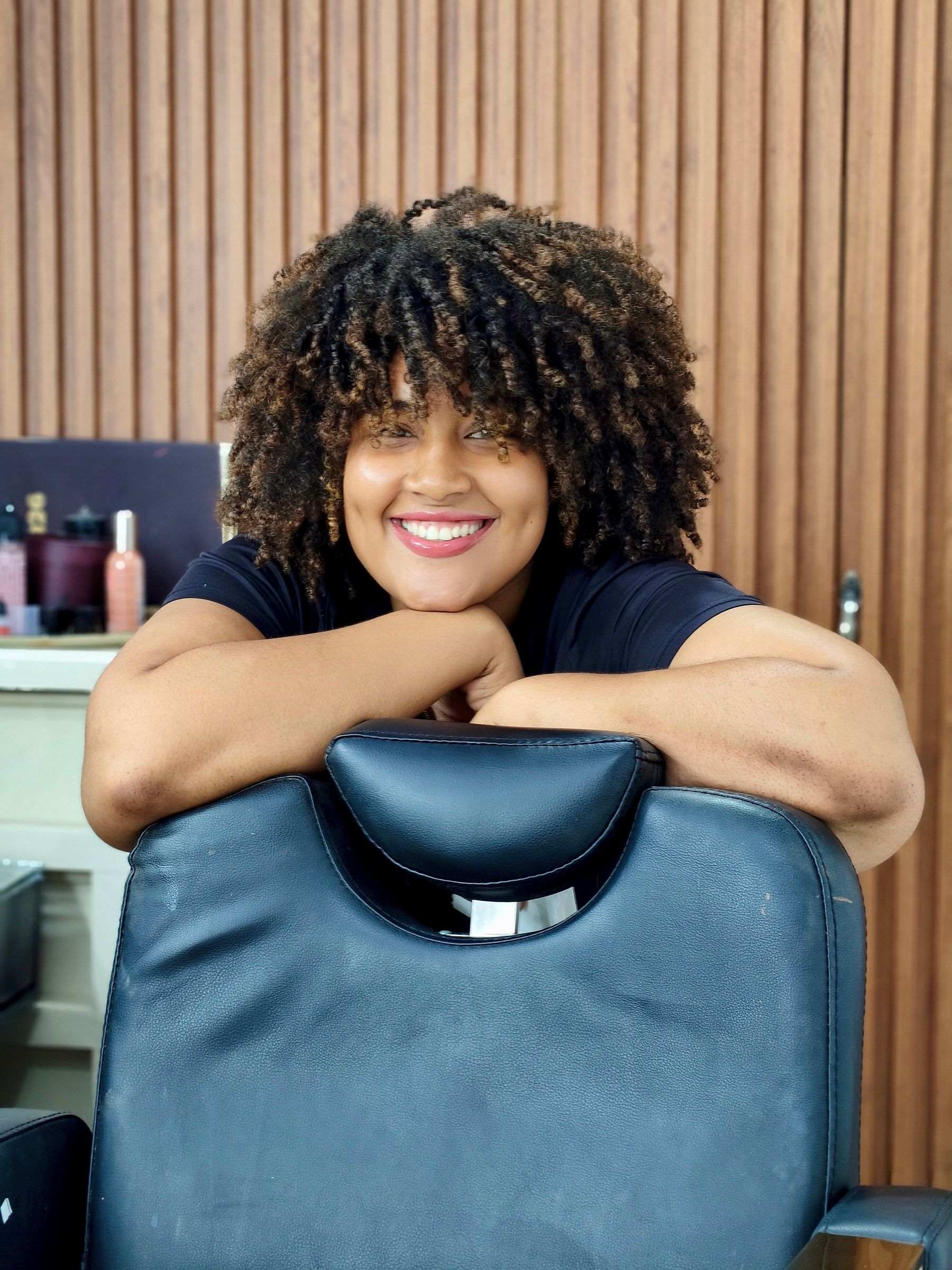 A smiling client with a curly afro leaning on the back of a salon chair, timber-slatted wall behind
